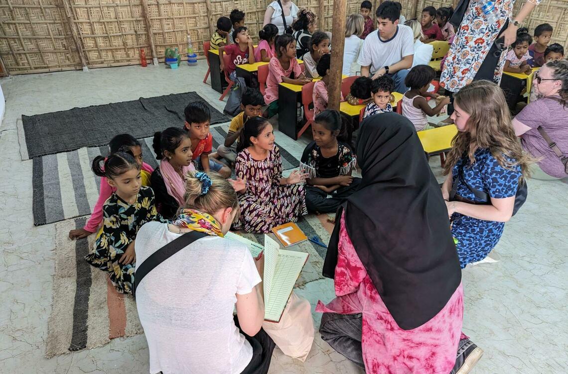 Students work with a local woman and children, crouched on the floor writing as they talk