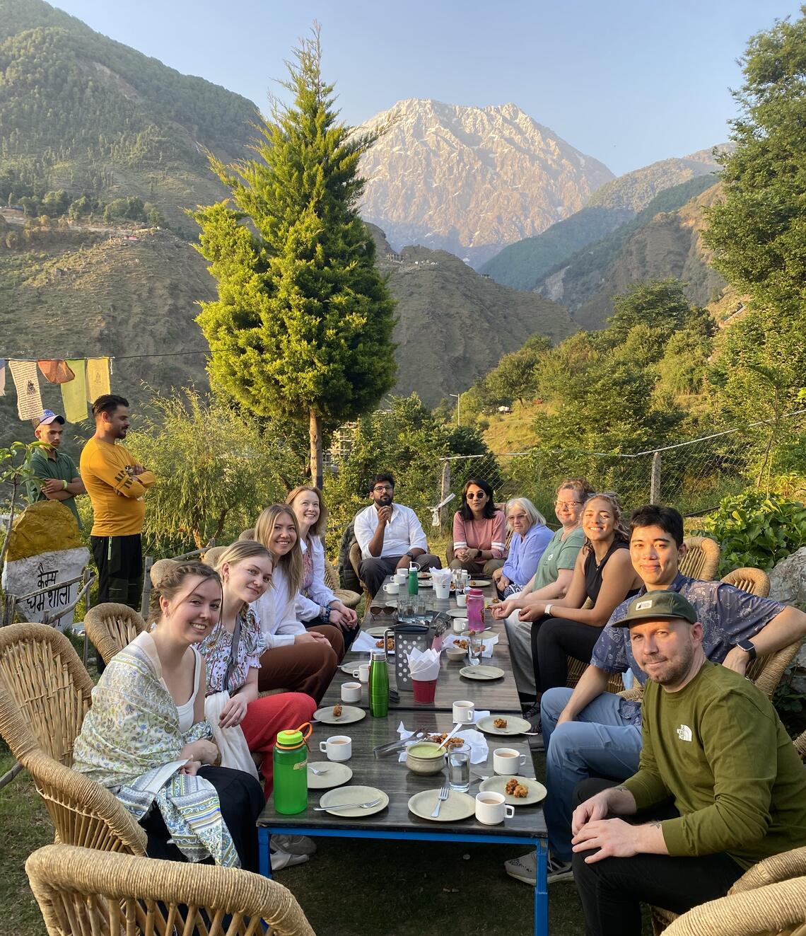 Group photo of students sitting around an outdoor dining table, having a meal under blue sky with a mountain in the background and Tibetan style prayer flags hanging from the side