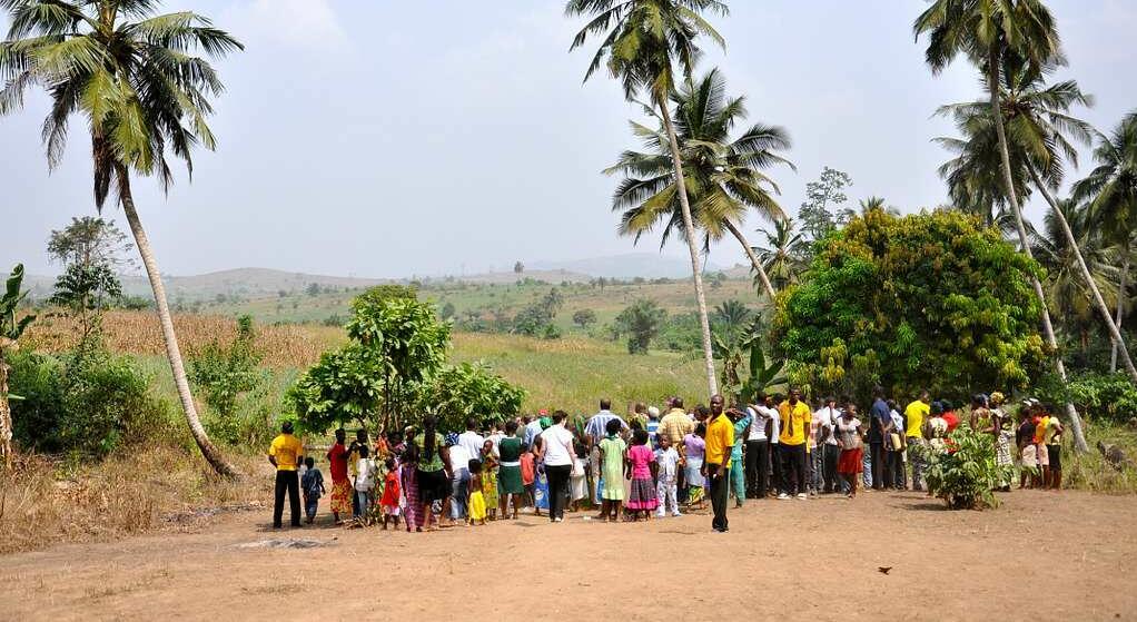 Colourfully dressed people, lined up at a water station outside a Ghanaian village with palm trees overhead