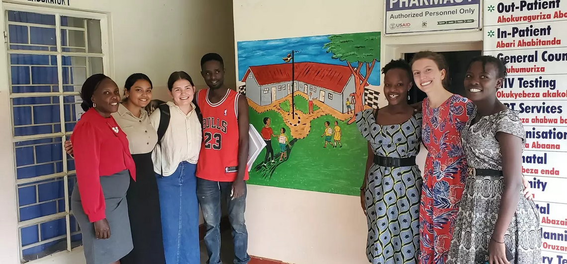 3 UCalgary students stand with 4 Ugandan students, posing in front of a hospital wall with a mural painting