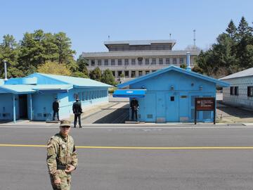 The foreground features a, presumably American, man in military uniform staring straight ahead(to the camera). The background features bright blue army base housing.