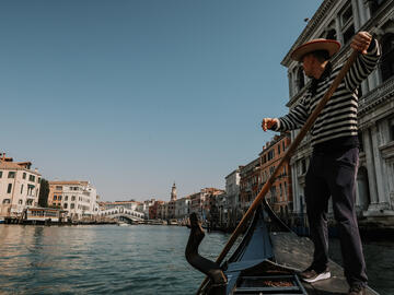 This photo is taken from a passenger point of view of a row boat at the Venice Grand Canal. The boatman stands at the edge of the boat, dressed in stripes and a hat, confidently looking onwards. The bliue waters mirror the blue skies and the venetian buildings frame either side of the canal.
