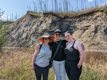 three women standing by a river with an embankment in the background on a sunny day