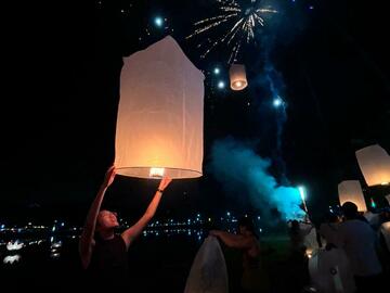 Hands lifting candle lanterns while more lanterns float in the sky above