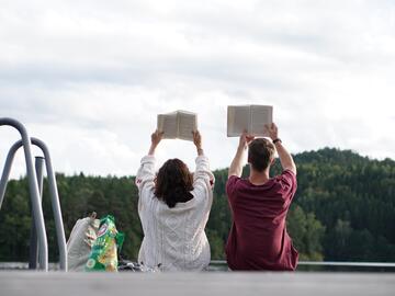Two people with their backs to the camera, sitting on a dock opposite a forest, holding books over their heads