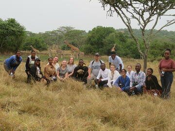 UCalgary and MUST students crouch in the grass for a photo on a safari tour, with giraffes walking far in the background