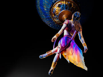 Performer dressed in purple and orange hanging from an aerial devise