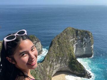Student looking back at the camera, smiling, while perched on a cliff with the ocean behind them