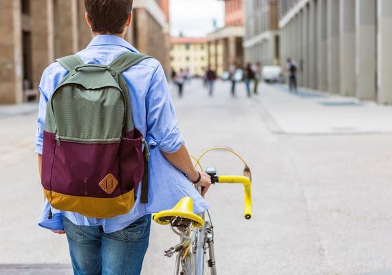 A short-haired person with a backpack stands beside a yellow bicycle, looking down a European-like street