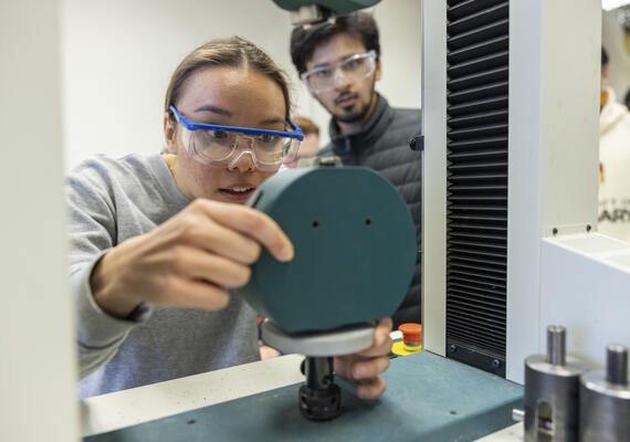 A student wearing safety glasses examines a piece of equipment in a lab.