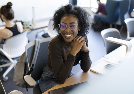 A young Black woman smiles sitting at a communal desk.