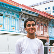 A man with dark skin & short hair stands with his hands behind his back. He smiles at the camera. His white shirt matches the white and teal framed buildings behind him.