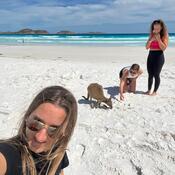 Selfie of a woman with long blonde hair. The camera is angled to show two other women behind her; one looks at a kangaroo while the other crouches with her hand out towards it. They are standing on a white sandy beach with blue-grey cloudy skies overhead.