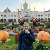A woman stands in the middle of a pumpkin field, with a castle-like building in the background. Her hair is tied back and she is taking a big bite out of a Danish hotdog.