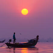 A silhouetted figure stands on a thin boat against a deep purple sky with a golden sun