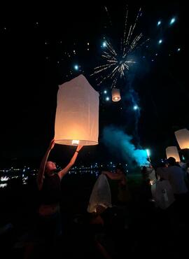 A crowd gathers in pitch dark. A woman's face is lit as she holds over her head a large paper lantern. Other lanterns drift around her into the night sky, along with a burst of fireworks.