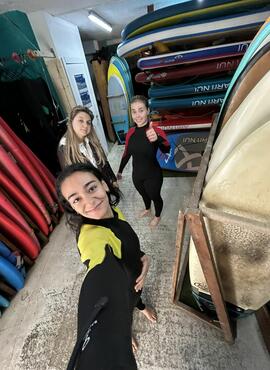 Three students in wetsuits smiling in front of surf boards