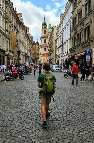 A man wearing a backpack walks down a cobblestone street with his back to the camera. Both sides of the road are framed by tall stone buildings and crowds of people. A church steeple in the distance is set against a blue sky.