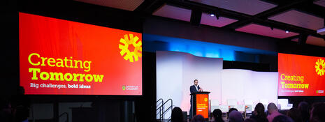 Person on a stage speaking to a large crowd flanked by giant screens