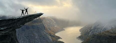 Two small silhouetted people, jumping with arms outstretched on top of a large cliff overhanging a fjord, lit by faint golden light through the clouds