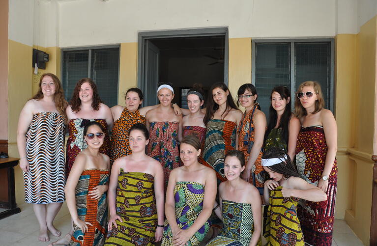 Group photo of 13 female students, wrapped in Ghanaian textile dresses and smiling for the camera