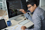 A young male sitting at a desk, pointing with a pen at a computer screen with code on it while he is looking at his notes beside him