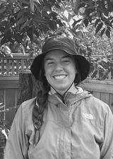 A happy young woman with long braided hair and a bucket hat stands in front of a lattice topped fence