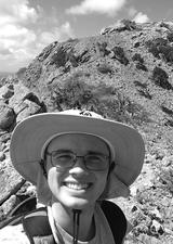 Joel Padgett standing on a hillside in the field with a wide brimmed hat and backpack