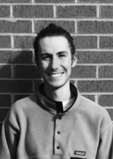 A happy young man with sharp features and hair in a bun stands in front of a brick wall