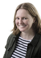 woman with strawberry blonde shoulder-length hair in a green jacket and striped blue and white shirt in front of a white background