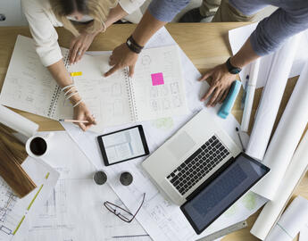 A top down view of a working table filled with paper and laptops and people sitting around the table.