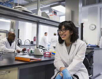 A student sits wearing a lab coat inside a lab.