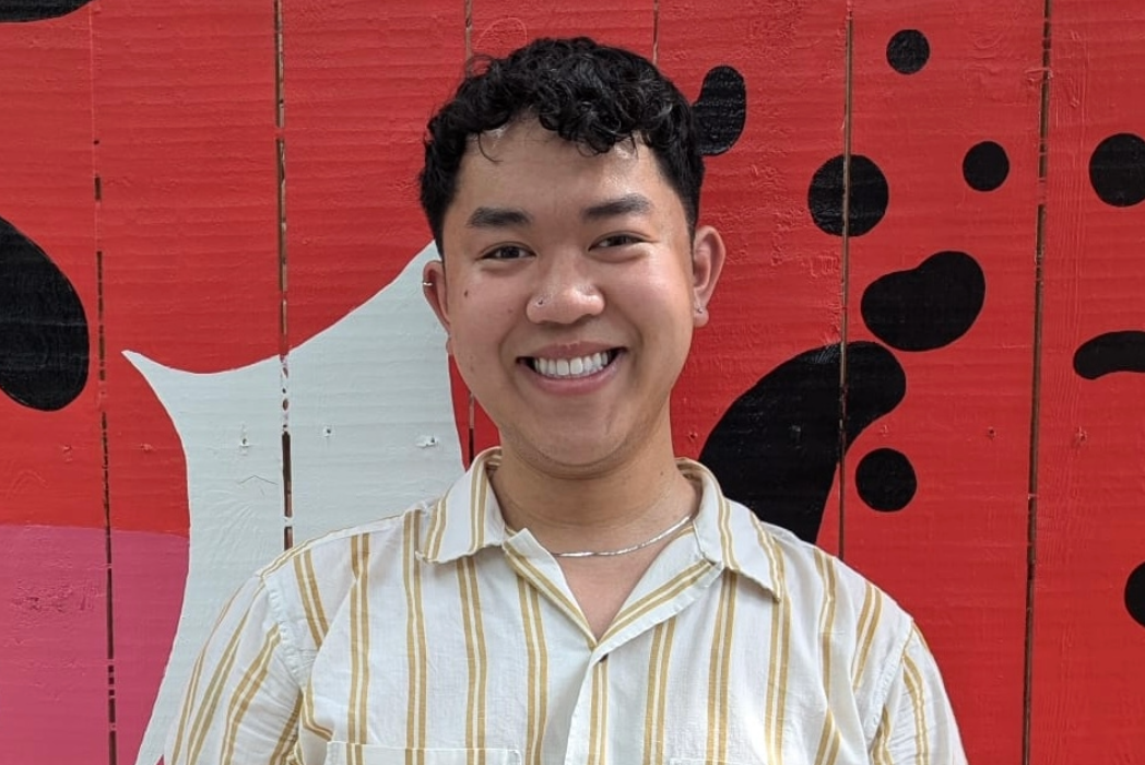 A man with short dark hair stands in front of a red wall